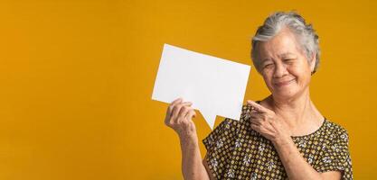 Concept of a paper speech bubble. An elderly Asian woman with short gray hair holding looking and pointing at a blank paper with a smile while standing on a yellow background in the studio photo