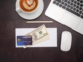Top view of a mockup credit card over on dollar banknote on white paper or envelope and a pencil with a laptop, mouse, and a coffee cup on the desk in the office. Vintage background photo