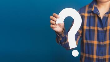 Hand of a businessman holding a white question mark paper while standing with a blue background. Close-up photo. Space for text photo
