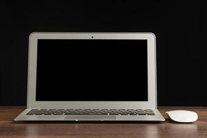 Laptop with blank screen and mouse on a wooden table with a black background. Space for text. Computer and technology concept photo