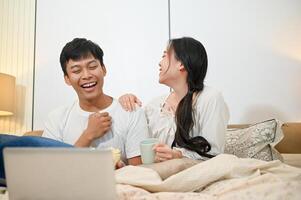 A cheerful Asian couple is laughing, enjoying watching a movie on a laptop in bed together. photo