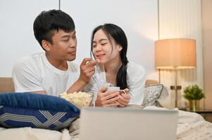 Happy Asian couple is having a fun time in bed, enjoying popcorn while watching a movie on a laptop. photo