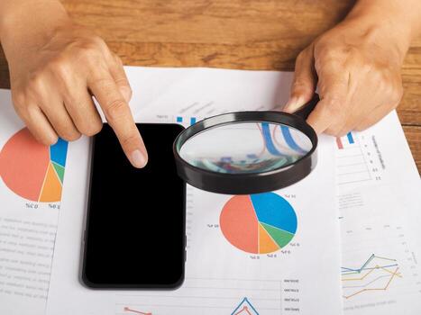 Hand of holding a magnifying glass reading financial data report and using a smartphone while sitting in the office. Close-up photo. Business and technology concept photo