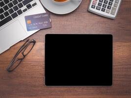 Top view of a tablet with a laptop, credit card, glasses, a coffee cup, and calculator on wooden table in the office. Flat lay, space for text. Working desk table concept photo