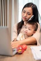 A busy mom is talking on the phone, focusing on her laptop, and taking care of her little son. photo
