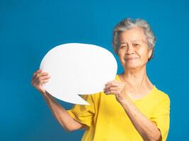 Concept of a speech bubble. Happy senior woman with short gray hair holding a blank paper and looking at the camera with a smile while standing on a blue background in the studio photo
