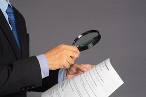 Businessman in a suit holding a magnifying glass and reading a document while standing over a gray background with copy space for text. Business and healthcare concept photo