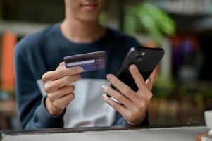 A cropped image of a positive young Asian man holding a smartphone and a credit card over a table. photo