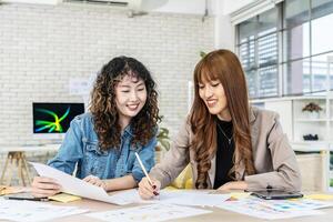 Two business young asian woman working together in office with reports and tablet, colleagues discuss financial data analyzing documents marketing project result talk business conversation photo