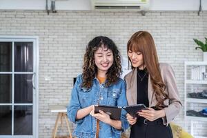 Portrait of cheerful two asian business woman standing in office room discussing collaborative project by using tablet. specifies details of task, talk to mentor at workspace.Teamwork photo
