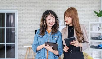 Portrait of cheerful two asian business woman standing in office room discussing collaborative project by using tablet. specifies details of task, talk to mentor at workspace.Teamwork photo