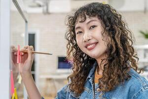 Young smiley asian creative business women are writing note on the text sheet after thinking new idea and planning project in glass wall, writing strategy business plan to development grow to success photo