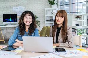 Two business young asian woman working in computer laptop together in office with reports, colleagues discuss financial data analyzing documents marketing project result talk business conversation photo
