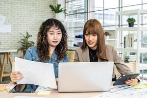 Two business young asian woman working in computer laptop together in office with reports, colleagues discuss financial data analyzing documents marketing project result talk business conversation photo