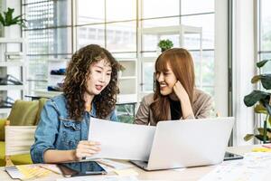 Two business young asian woman working in computer laptop together in office with reports, colleagues discuss financial data analyzing documents marketing project result talk business conversation photo