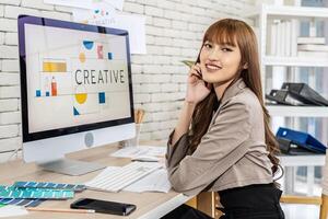 Asian young woman graphic designer smiling and looking at camera sitting at the desk with pc and tablet computer while working in office, Artist Creative Designer Illustrator Graphic Skill Concept photo