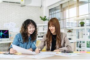 Two business young asian woman working together in office with reports and tablet, colleagues discuss financial data analyzing documents marketing project result talk business conversation photo