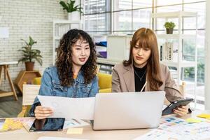 Two business young asian woman working in computer laptop together in office with reports, colleagues discuss financial data analyzing documents marketing project result talk business conversation photo