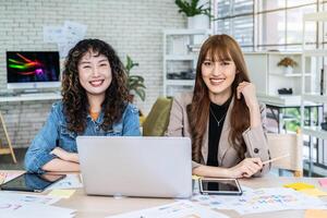 Two business young asian woman working in computer laptop together in office with reports, colleagues discuss financial data analyzing documents marketing project result talk business conversation photo