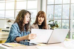 Two business young asian woman working in computer laptop together in office with reports, colleagues discuss financial data analyzing documents marketing project result talk business conversation photo