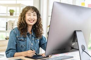 Asian young woman graphic designer smiling and looking at camera sitting at the desk with pc and tablet computer while working in office, Artist Creative Designer Illustrator Graphic Skill Concept photo