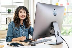 Asian young woman graphic designer smiling and looking at camera sitting at the desk with pc and tablet computer while working in office, Artist Creative Designer Illustrator Graphic Skill Concept photo