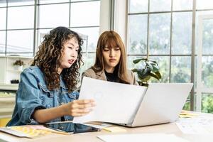 Two business young asian woman working in computer laptop together in office with reports, colleagues discuss financial data analyzing documents marketing project result talk business conversation photo