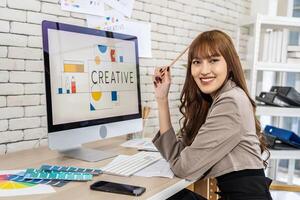 Asian young woman graphic designer smiling and looking at camera sitting at the desk with pc and tablet computer while working in office, Artist Creative Designer Illustrator Graphic Skill Concept photo