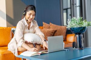 Consultant girl wearing headset with microphone sitting on sofa,, freelance worker, call center or support service operator helpline. Young woman having talk with client or colleague and take notes photo