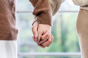 Close up of hand young happy loving couple guy and girl standing in front of big window, back to camera front view of an affectionate couple looking out of window while holding hand photo