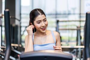 Young asian athletic woman with perfect shape body wearing earphones is choosing workout music with smart phone to listen while practicing in gym, Technology to stay fit and motivated photo
