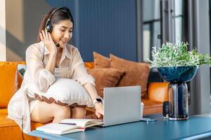 Consultant girl wearing headset with microphone sitting on sofa,, freelance worker, call center or support service operator helpline. Young woman having talk with client or colleague and take notes photo