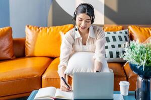 Consultant girl wearing headset with microphone sitting on sofa,, freelance worker, call center or support service operator helpline. Young woman having talk with client or colleague and take notes photo