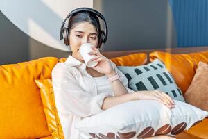 Weekend relax,Young asian Woman In Headphones Sitting In Couch At Home Listening Favorite Music and enjoying cup of coffee on sofa, Concept Of Relax And Meditation photo