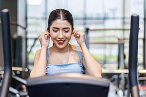 Young asian athletic woman with perfect shape body wearing earphones is choosing workout music with smart phone to listen while practicing in gym, Technology to stay fit and motivated photo