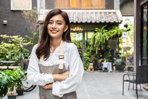 Successful small business young asian owner woman standing with looking at camera posing crossing arms, Portrait of waiter salesman worker wearing apron standing at work space smiling friendly photo