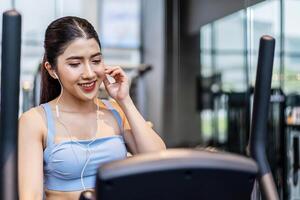 Young asian athletic woman with perfect shape body wearing earphones is choosing workout music with smart phone to listen while practicing in gym, Technology to stay fit and motivated photo