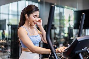 Young asian athletic woman with perfect shape body wearing earphones is choosing workout music with smart phone to listen while practicing in gym, Technology to stay fit and motivated photo