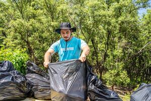 One volunteers in work overalls collect garbage in forest in plastic bag. Close-up during action. Cleaning of nature from waste. Environmental assistance. Garbage collection in forest photo