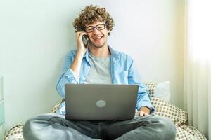 Caucasian Freelancer Guy Talking On Phone Using Laptop Computer Communicating Online Working on the bed in the room, Distance Work And Mobile Communication Concept. photo