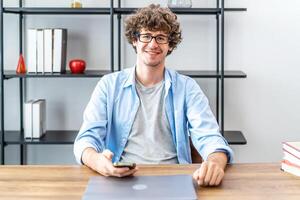 Young and successful. Smiling business man wearing eyeglasses holding mobile phone and looking at camera while sitting at desk in home, Caucasian businessman using smartphone photo
