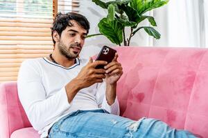 Cool gadget and app. Young smiling guy holding mobile phone, typing sms message, resting on sofa in living room, Texting And Browsing Internet, surfing web, using app, Networking In Social Media photo