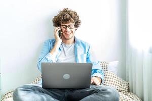 Caucasian Freelancer Guy Talking On Phone Using Laptop Computer Communicating Online Working on the bed in the room, Distance Work And Mobile Communication Concept. photo