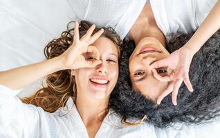 Top view self portrait of caucasian and black women wearing sleep with happy face smiling doing ok sign with hand on eye looking through fingers, looking at camera photo