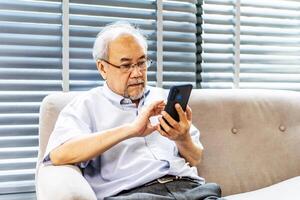 Pleasant senior older man resting on couch at home, older middle aged male customer holding smartphone using mobile app, texting message, search ecommerce offers on cell phone technology device photo