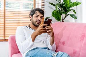 Cool gadget and app. Young smiling guy holding mobile phone, typing sms message, resting on sofa in living room, Texting And Browsing Internet, surfing web, using app, Networking In Social Media photo