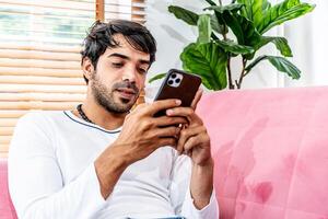 Cool gadget and app. Young smiling guy holding mobile phone, typing sms message, resting on sofa in living room, Texting And Browsing Internet, surfing web, using app, Networking In Social Media photo