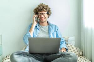 Caucasian Freelancer Guy Talking On Phone Using Laptop Computer Communicating Online Working on the bed in the room, Distance Work And Mobile Communication Concept. photo