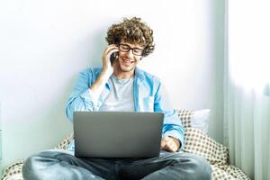 Caucasian Freelancer Guy Talking On Phone Using Laptop Computer Communicating Online Working on the bed in the room, Distance Work And Mobile Communication Concept. photo