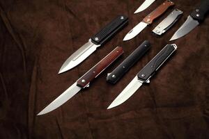 A variety of folding and pocket knives lie on brown fabric. A versatile pocket tool and self-defense tool. photo
